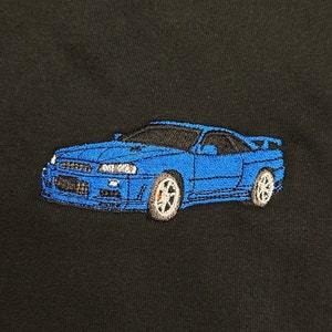 May include: Embroidered blue sports car on a black background. The car features detailed stitching, including headlights, wheels, and a spoiler. The car's body is a vibrant blue, contrasting with the black fabric.