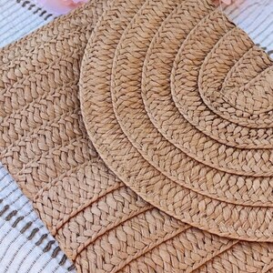 May include: Close-up of woven placemats in a natural tan color. The placemats have a textured, braided design. The image shows a set of round and rectangular placemats, perfect for a dining table setting. A peach-colored flower is in the background.