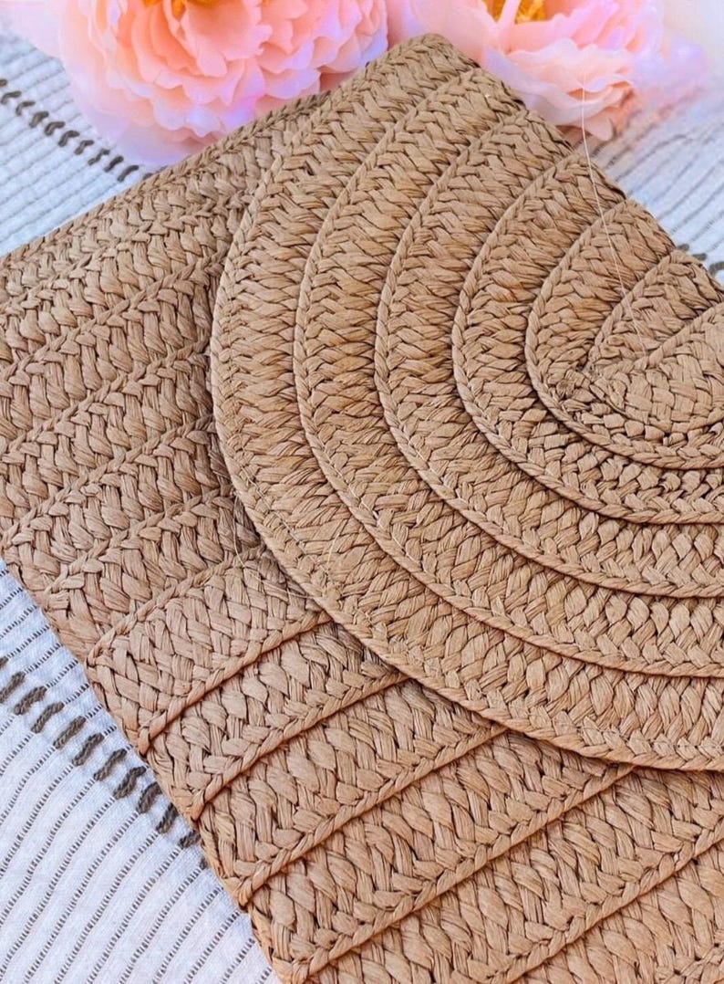 May include: Close-up of woven placemats in a natural tan color. The placemats have a textured, braided design. The image shows a set of round and rectangular placemats, perfect for a dining table setting. A peach-colored flower is in the background.
