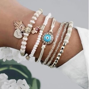 Könnte beinhalten: Sechs Armbänder mit weißen Perlen, goldenen Akzenten und Charms. Die Charms sind eine Hamsa-Hand, ein böses Auge und ein Herz mit dem Wort "love" darauf.