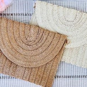 May include: Two woven straw clutch purses in natural and cream colors. The purses have a flap closure and a textured, fan-like design on the front. The background is a striped fabric with pink flowers.