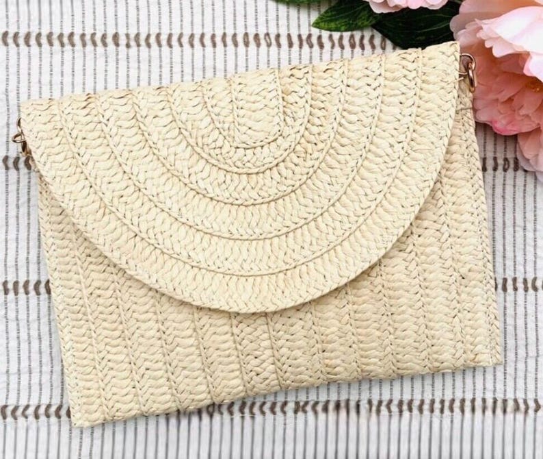 May include: A woven straw clutch with a flap closure. The clutch is a light beige color and has a textured surface. The flap has a semi-circular design. The clutch has a gold-colored chain strap.