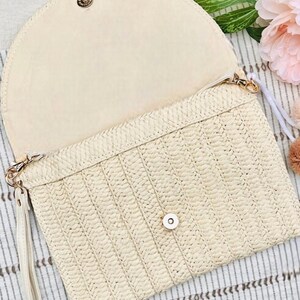 May include: Open, cream-colored clutch with a woven straw exterior and a matching interior. The clutch features a flap closure with a gold-tone button and a wrist strap. The bag is designed with a textured pattern.