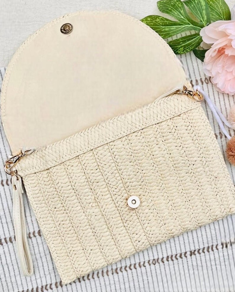 May include: Open, cream-colored clutch with a woven straw exterior and a matching interior. The clutch features a flap closure with a gold-tone button and a wrist strap. The bag is designed with a textured pattern.
