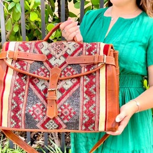 May include: A rectangular satchel bag with a woven textile front featuring a geometric pattern in red, black, and cream. The bag has brown leather trim, straps, and a buckle closure. It has a top handle and a shoulder strap.