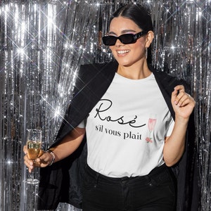May include: A white t-shirt with the text "Rosè s'il vous plaît" and a pink illustration of a glass of rosé wine. The person wearing the shirt is holding a glass of champagne.