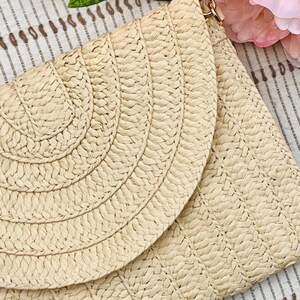 May include: A beige woven clutch with a flap closure. The clutch features a geometric pattern on the body and a curved design on the flap. A gold-colored clasp is visible at the top. Pink flowers are in the background.