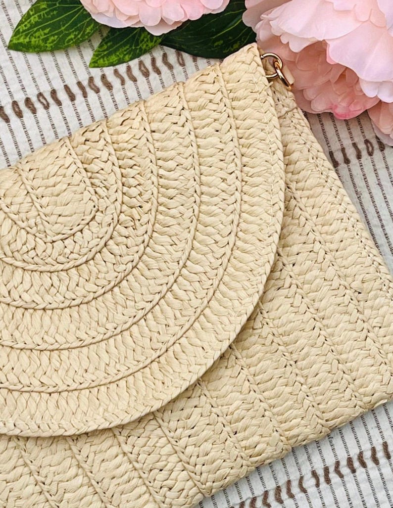 May include: A beige woven clutch with a flap closure. The clutch features a geometric pattern on the body and a curved design on the flap. A gold-colored clasp is visible at the top. Pink flowers are in the background.