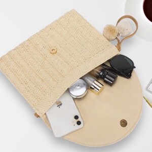 May include: A beige straw clutch bag with a flap closure, revealing a cream-colored interior. The bag contains a smartphone, sunglasses, and cosmetic items. A small handle with pom-poms is attached. The bag is open, showing its contents.