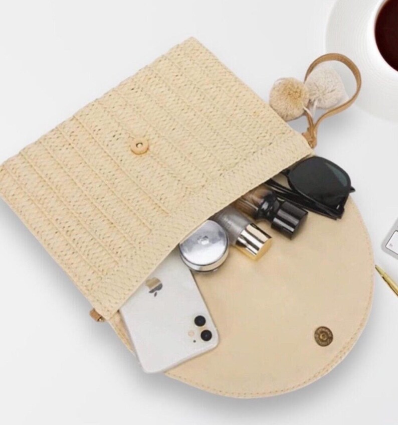 May include: A beige straw clutch bag with a flap closure, revealing a cream-colored interior. The bag contains a smartphone, sunglasses, and cosmetic items. A small handle with pom-poms is attached. The bag is open, showing its contents.