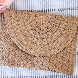 May include: A rectangular, woven straw clutch with a rounded flap. The clutch is tan with a woven pattern and a gold-colored metal ring on the side. The flap has a semi-circular design. The background is a striped fabric.