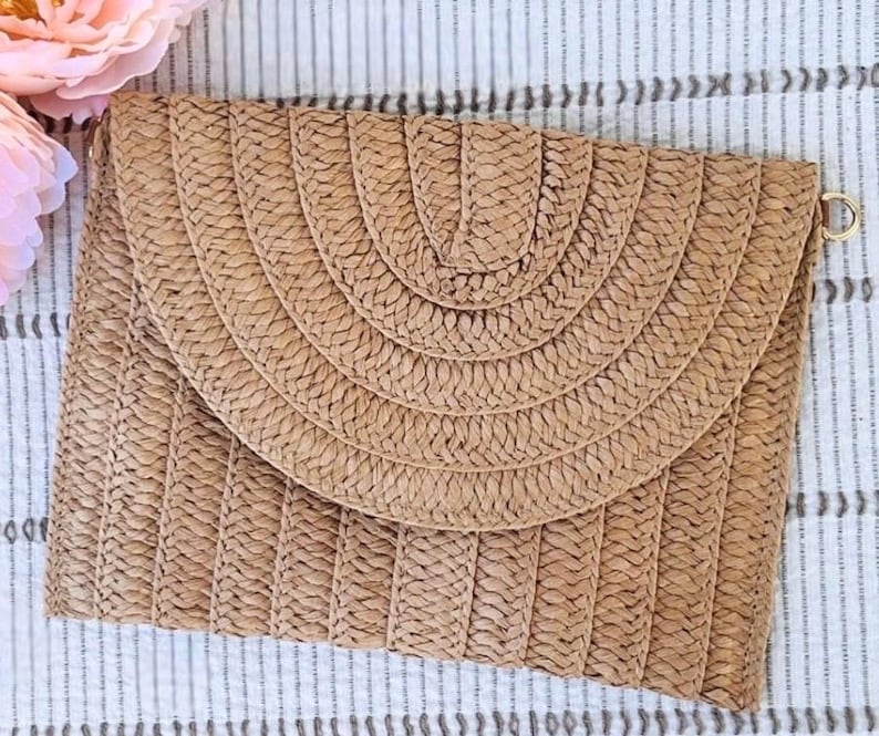 May include: A rectangular, woven straw clutch with a rounded flap. The clutch is tan with a woven pattern and a gold-colored metal ring on the side. The flap has a semi-circular design. The background is a striped fabric.