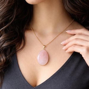 May include: A gold chain necklace featuring a large, pink, teardrop-shaped gemstone pendant with a gold border. The necklace is worn over a dark top. The pendant is a soft pink colour.