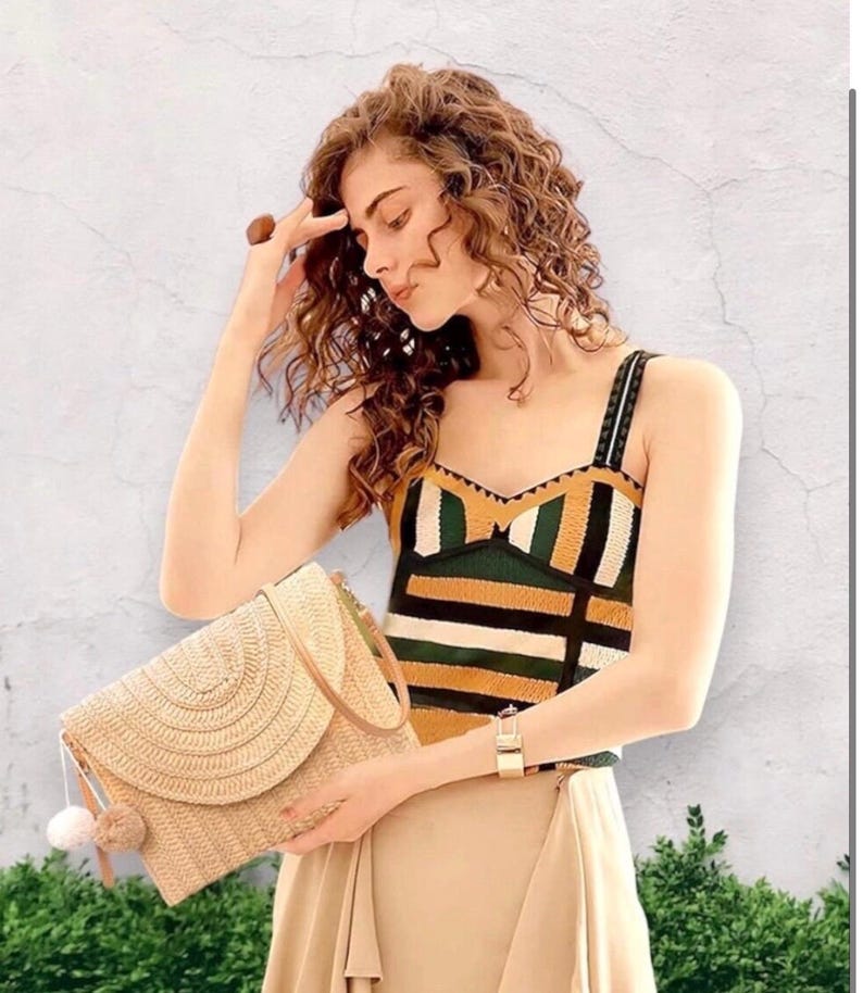 May include: A straw clutch with a flap closure and a beige pom-pom tassel. The clutch is held in front of a person wearing a patterned top with green, black, and yellow stripes. A gold watch is visible on the wrist.