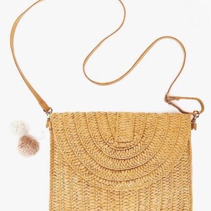 May include: A woven straw crossbody bag with a long, thin, tan strap. The bag has a flap closure with a semi-circular design. Two small pom-poms hang from the side. The bag is a light tan color.