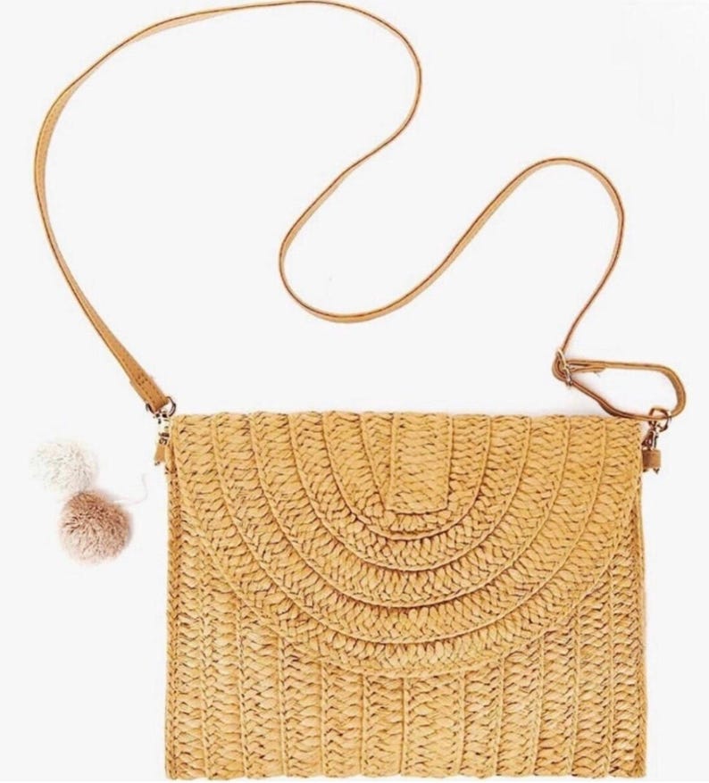 May include: A woven straw crossbody bag with a long, thin, tan strap. The bag has a flap closure with a semi-circular design. Two small pom-poms hang from the side. The bag is a light tan color.