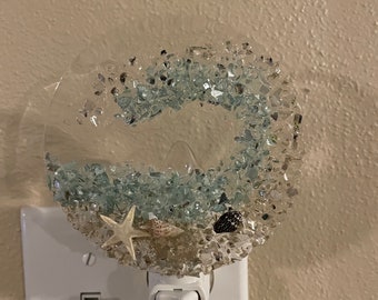 May include: A clear glass nightlight plug-in with a blue and white wave design and seashells. The nightlight is plugged into a white electrical outlet.