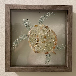 May include: A framed mosaic art piece depicting a sea turtle made from iridescent glass and shell pieces. The turtle is in a light brown wooden frame with a glass front.