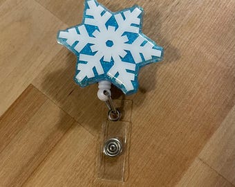May include: A blue snowflake-shaped badge holder with glitter and a white snowflake design. The holder has a clear plastic retractable cord and a clear plastic clip.