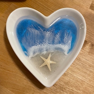 May include: A white heart-shaped dish with a blue and white resin design resembling an ocean scene. A white starfish is in the center of the dish.