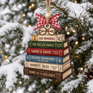 May include: A wooden Christmas tree ornament shaped like a stack of six books with a red and white patterned bow and glasses on top. The book spines are labeled 2026 OUR MEMORIES, THE MILLER FAMILY FESTIVE TALES, SARAH & DAVID WINTER STORIES, HOLIDAY ADVENTURES THE MILLERS, FAVORITE TALES CHRISTMAS 2026, and HOME FOR THE HOLIDAYS.