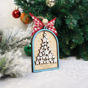 May include: A wooden Christmas tree ornament with a light blue border and a natural wood interior. The ornament features a star-shaped tree with names and the text "MRS. KRUEGER 2024-2025". It is adorned with a red and white bow.