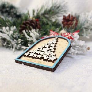 May include: Wooden Christmas tree ornament with a light blue border. The ornament features a tree made of star-shaped cutouts with names on them. The text "MRS. KRUEGER 2024-2025" is at the bottom. The background includes pine branches and pine cones.