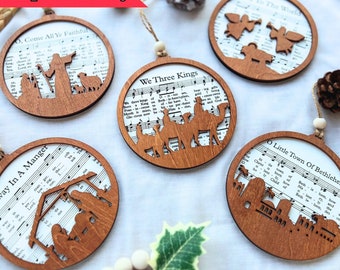 May include: Wooden Christmas ornaments with religious themes. Each round ornament features a cutout design of a nativity scene or angels, set against a backdrop of sheet music. The ornaments are strung with twine.