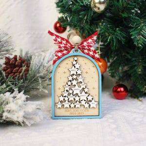 May include: A wooden Christmas ornament shaped like a tree, with a light blue frame and a red and white bow. The ornament features star cutouts with names and the text "MRS. KRUEGER 2024-2025".