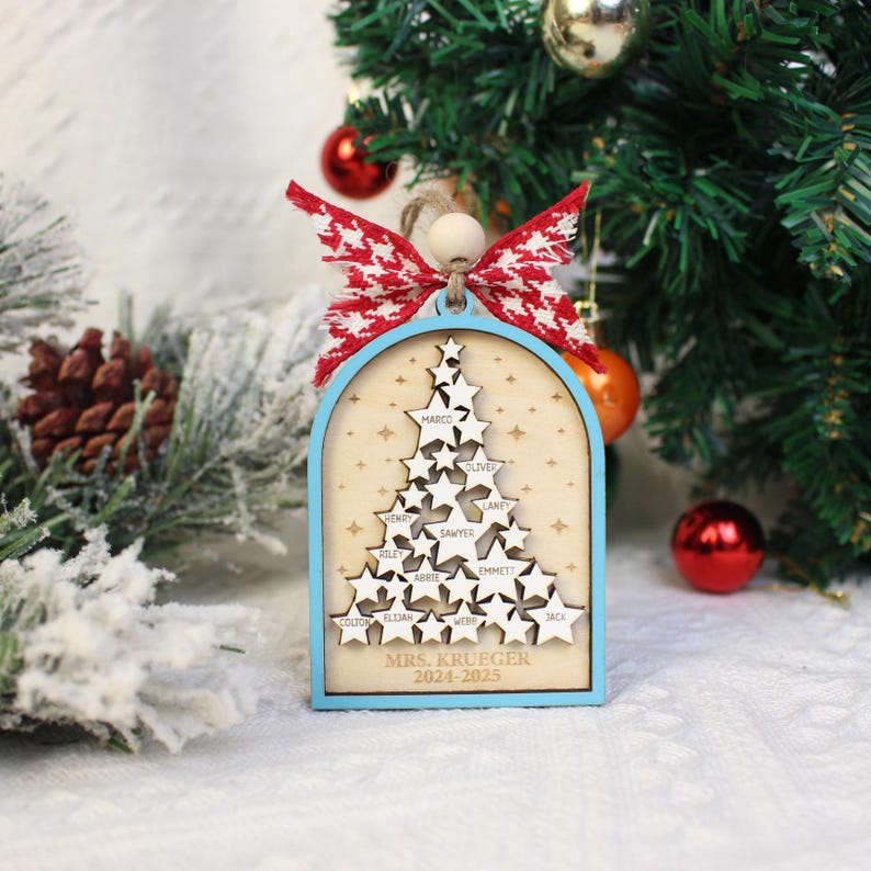 May include: A wooden Christmas ornament shaped like a tree, with a light blue frame and a red and white bow. The ornament features star cutouts with names and the text "MRS. KRUEGER 2024-2025".