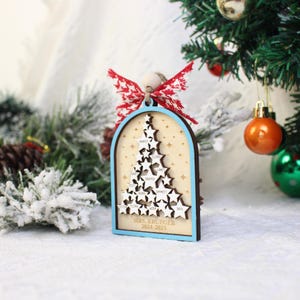 May include: A wooden Christmas tree ornament with a light blue border and a star-shaped tree design. The ornament features names and the text "MRS. KRUEGER 2024-2025". It is adorned with a red and white bow and is hanging near a Christmas tree.