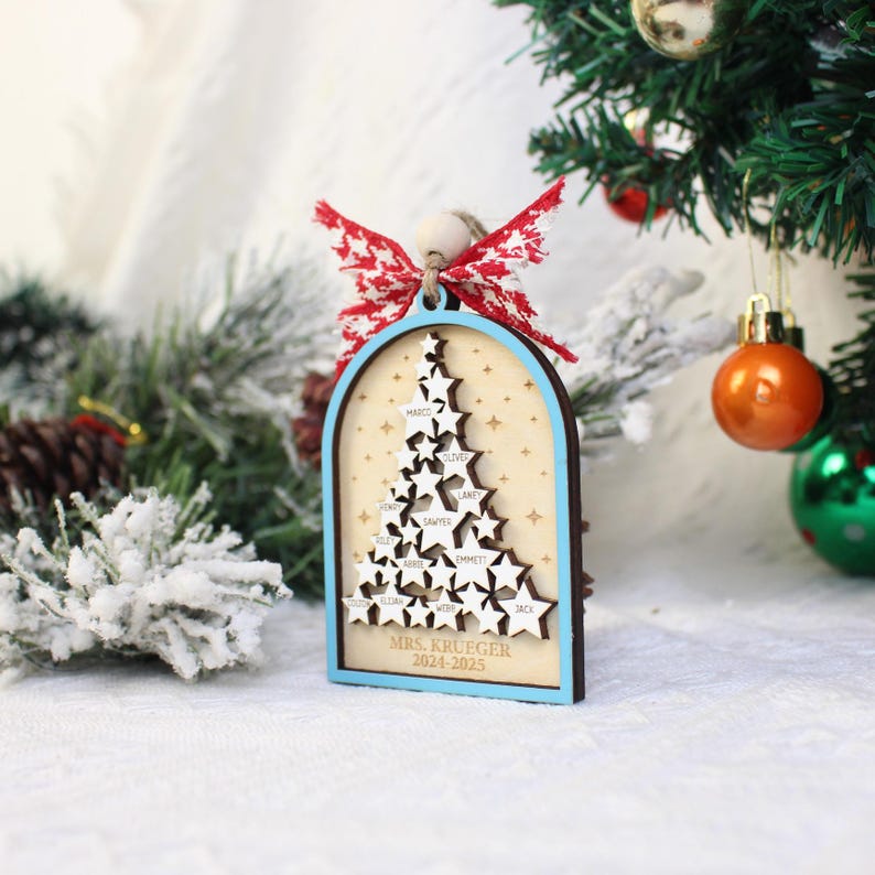 May include: A wooden Christmas tree ornament with a light blue border and a star-shaped tree design. The ornament features names and the text "MRS. KRUEGER 2024-2025". It is adorned with a red and white bow and is hanging near a Christmas tree.