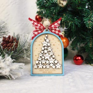May include: A wooden Christmas tree ornament with a light blue frame and a red and white bow. The ornament features a tree made of star cutouts with names and the text "MRS. KRUEGER 2024-2025".