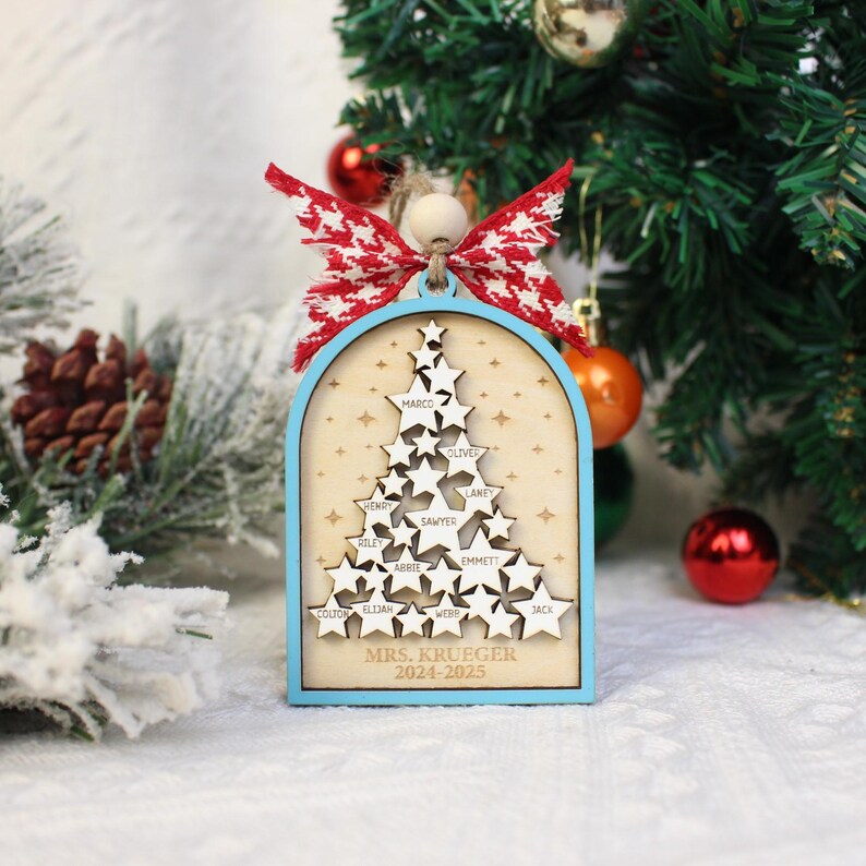 May include: A wooden Christmas tree ornament with a light blue frame and a red and white bow. The ornament features a tree made of star cutouts with names and the text "MRS. KRUEGER 2024-2025".