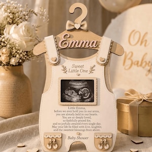 May include: A wooden baby onesie-shaped plaque featuring a hanger top with a bow, the name Emma, and a central frame for an ultrasound photo. It includes the text Sweet Little One, a poem about being held in hearts, and Baby Shower with small shoe icons.