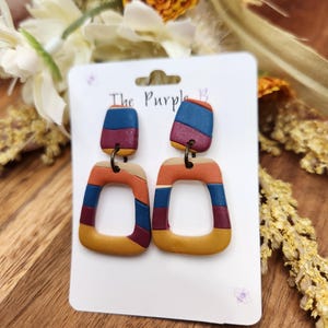 May include: A pair of colorful, geometric earrings. Each earring features a rectangular top and a square-shaped bottom, with a repeating pattern of blue, orange, and maroon stripes. The earrings are attached to a white card with the text "The Purple B".