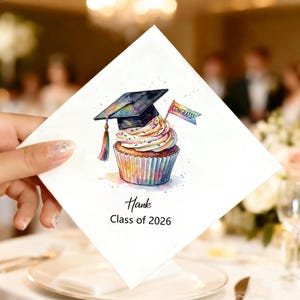 May include: White square napkin with a watercolor illustration of a graduation cupcake. The cupcake has a graduation cap, a rainbow-colored frosting, and a small flag that says "Congrats!". The napkin also has the name "Hank" and "Class of 2026" printed on it.