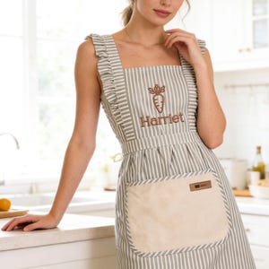 May include: A gray and white striped apron with a large front pocket and ruffle trim. The apron features an embroidered carrot design and the name "Harriet". The apron is made of a light-colored fabric.