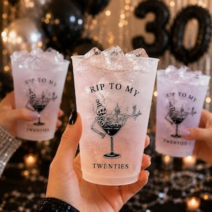 May include: A clear plastic party cup filled with ice and a light pink beverage. The cup features a black graphic of a skeleton wearing a party hat inside a martini glass, with the text RIP TO MY TWENTIES printed in black lettering.