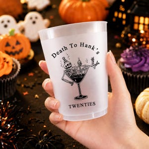 May include: A clear plastic party cup featuring a black graphic of a skeleton wearing a party hat inside a martini glass. The text "Death To Hank's" is printed above the skeleton, and the word "TWENTIES" is printed below the glass.