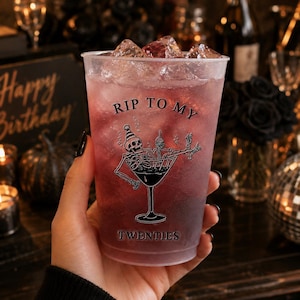 May include: A clear plastic cold cup filled with a dark purple beverage and ice cubes. The cup features a black graphic of a skeleton wearing a party hat inside a martini glass, with the text RIP TO MY TWENTIES printed in black lettering.