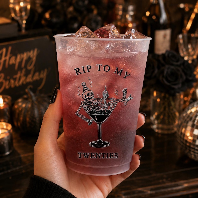 May include: A clear plastic cold cup filled with a dark purple beverage and ice cubes. The cup features a black graphic of a skeleton wearing a party hat inside a martini glass, with the text RIP TO MY TWENTIES printed in black lettering.