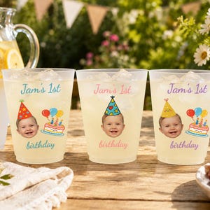 Könnte beinhalten: Drei durchsichtige Kunststoff-Partybecher, gefüllt mit Eis und einem hellen Getränk. Jeder Becher zeigt einen Aufdruck eines Babys mit buntem Partyhut, eine Geburtstagstorte mit Kerzen sowie den Schriftzug "Jam's 1st birthday" in verschiedenen Farben.