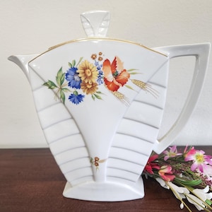 May include: A white porcelain teapot with a fan-shaped body and a geometric ribbed design. The front features a colorful floral bouquet of blue cornflowers, yellow daisies, and orange poppies with wheat stalks. It has a gold rim and lid finial.