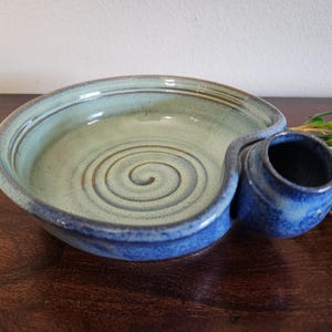 May include: Handmade ceramic serving bowl with a blue and green glaze. The bowl features a spiral design in the center and a small attached compartment. The rim and the compartment are a darker blue.