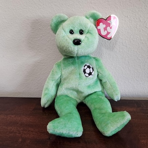 Puede incluir: Un oso de peluche verde con un emblema de balón de fútbol en el pecho. El oso tiene ojos negros y una nariz negra. Una etiqueta en forma de corazón está unida a su oreja.