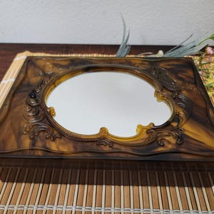 May include: A rectangular, tortoiseshell-patterned box with an oval mirror. The box features ornate, swirling details around the mirror and on the frame. The box is set on a bamboo mat.