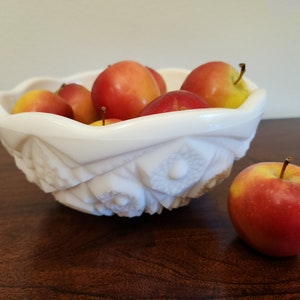 May include: A white, decorative bowl filled with red and yellow apples. The bowl has a scalloped rim and a detailed, embossed floral pattern. One apple sits beside the bowl. The apples are a mix of red and yellow.