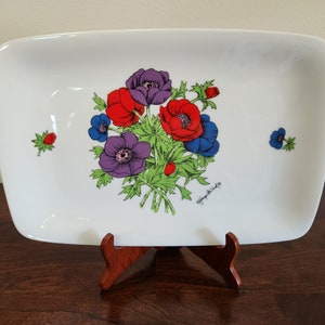 Puede incluir: Plato rectangular blanco con esquinas redondeadas, decorado con un ramo floral de anémonas rojas, moradas y azules. Las flores se colocan sobre un follaje verde. El plato se exhibe sobre un soporte de madera.