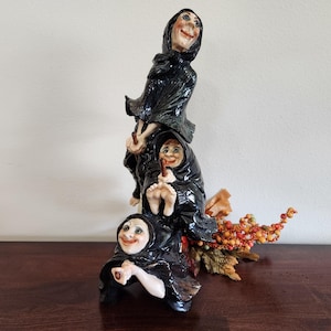 Op de afbeelding: Een grillige keramische sculptuur van drie lachende heksen in zwarte mantels. De heksen zijn op elkaar gestapeld en houden kleine stokjes vast. Naast de heksen staat een decoratief arrangement van bessen en bladeren.